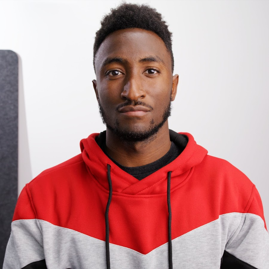 A head-and-shoulders portrait of Marques Brownlee, a young Black man with a close-cropped afro and a slight smile. He is wearing a red and gray hoodie and looking directly at the camera. The background is a solid dark gray.