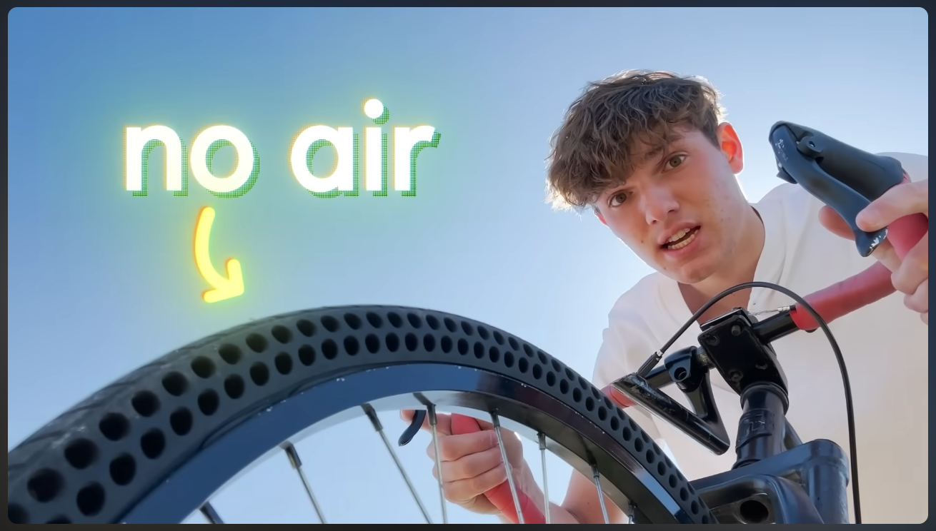 A close-up shot of a bicycle's front wheel and tire against a bright, slightly hazy blue sky. A young man with short brown hair, identified as Brandon Voloch, is visible from the chest up on the right side of the frame, looking intently at the tire. He is holding a black tool near the tire. The tire itself is black and features numerous circular perforations instead of an inflatable tube. The text "no air" is superimposed in large, glowing white letters at the top left, with a glowing yellow arrow pointing downwards towards the airless tire. The image emphasizes the unique, airless design of the bicycle tire.